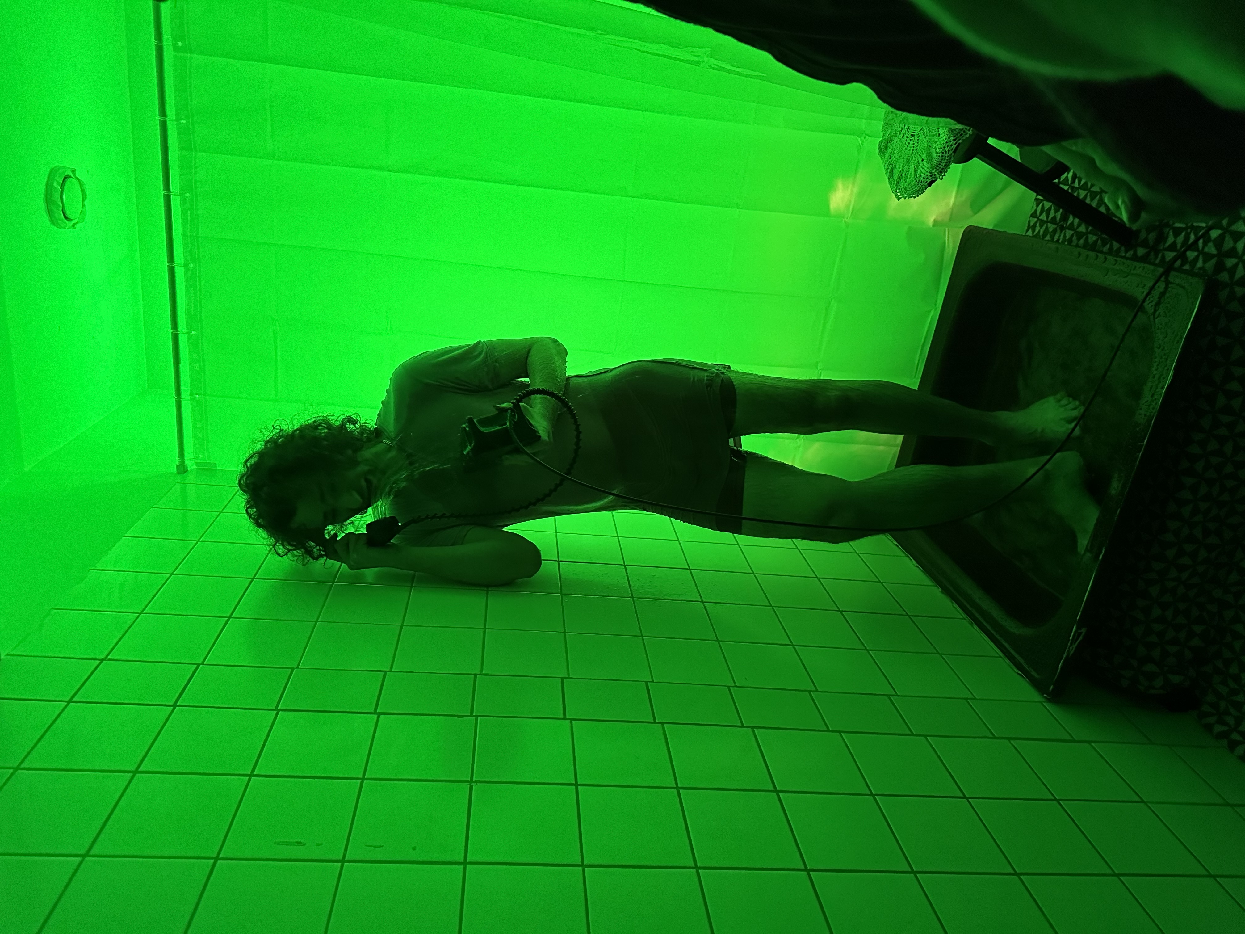 A tiled bathroom is washed in green light, silhouetting me standing in a shower, speaking into a rotary phone, which is spraying water